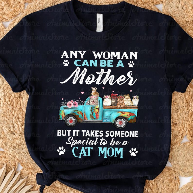 Puede incluir: Camiseta azul marino con el texto "ANY WOMAN CAN BE A MOTHER BUT IT TAKES SOMEONE SPECIAL TO BE A CAT MOM." Un cami&oacute;n turquesa transporta gatos y flores. La camiseta tiene un dise&ntilde;o desgastado.