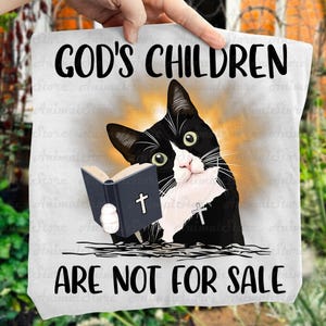 Puede incluir: Una bolsa de mano blanca con una ilustración de un gato blanco y negro sosteniendo un libro con una cruz. El texto "GOD'S CHILDREN ARE NOT FOR SALE" está impreso encima y debajo de la imagen del gato.
