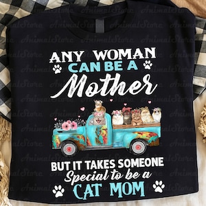 Puede incluir: Camiseta negra con el texto "ANY WOMAN CAN BE A MOTHER BUT IT TAKES SOMEONE SPECIAL TO BE A CAT MOM." Una caja de cami&oacute;n azul est&aacute; llena de gatos con coronas de flores. La camiseta tiene un fondo a cuadros.
