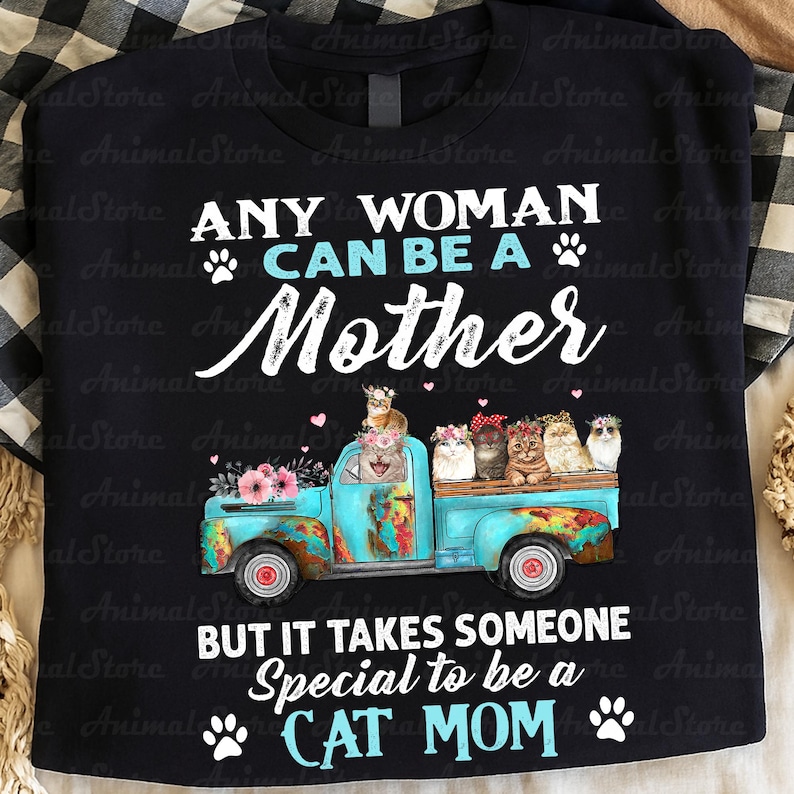 Puede incluir: Camiseta negra con el texto "ANY WOMAN CAN BE A MOTHER BUT IT TAKES SOMEONE SPECIAL TO BE A CAT MOM." Una caja de cami&oacute;n azul est&aacute; llena de gatos con coronas de flores. La camiseta tiene un fondo a cuadros.