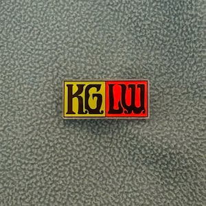 May include: A rectangular pin with the text "KG LUU" in yellow and red, set against a green fleece jacket. The jacket has an orange zipper and a textured surface.