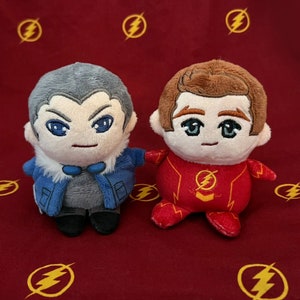 Puede incluir: Dos juguetes de peluche, uno con una chaqueta azul y gris y el otro con un traje rojo y amarillo. El juguete con la chaqueta azul tiene una cara blanca con ojos negros y una boca roja. El juguete con el traje rojo tiene una cara marrón con ojos negros y una boca roja. Ambos juguetes tienen un símbolo de rayo en el pecho.