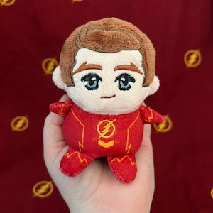 Puede incluir: Un muñeco de peluche de un personaje con un disfraz rojo y amarillo. El muñeco tiene cabello castaño y ojos grandes bordados. El disfraz presenta un emblema de rayo en el pecho. El fondo es una tela granate con símbolos de rayo amarillos.