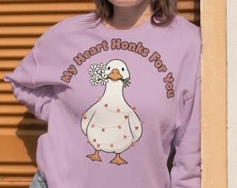 May include: A light purple sweatshirt with the text "My Heart Honks For You" in a circular design. The graphic features a white goose holding flowers, with a string of hearts. The sweatshirt is a long-sleeved design.