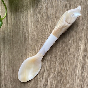 Może przedstawiać: Biała i brązowa łyżka z perłowej muszli z uchwytem w kształcie głowy ryby. Łyżka leży na drewnianej powierzchni.