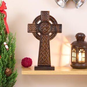 Może przedstawiać: Drewniany celtycki krzyż w ciemnobrązowym kolorze z misternymi wzorami węzłów. Krzyż stoi na drewnianej podstawie i jest wystawiony na półce z czerwoną kulą i latarnią.