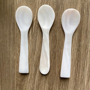 Puede incluir: Tres cucharas blancas de nácar sobre una superficie de madera. Las cucharas son pequeñas y tienen una forma redondeada.