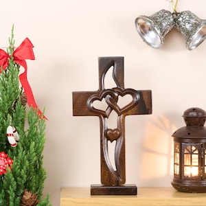 May include: A wooden cross with two intertwined hearts carved into the centre. The cross is brown and has a simple base. It is displayed on a wooden shelf with a small Christmas tree, a lantern, and silver bells.