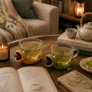 May include: A cozy scene featuring two clear glass teacups filled with tea, resting on handmade crochet frog coasters. A lit candle, teapot, and open book are also on the wooden table. The candle has the text "COZY MOMENT".