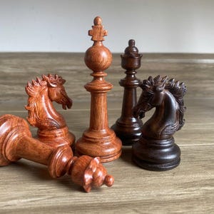 Könnte beinhalten: Eine Sammlung von fünf hölzernen Schachfiguren, bestehend aus zwei Springern, einem aufrecht stehenden König, einer Dame und einem auf der Seite liegenden König. Die Figuren sind aus zwei verschiedenen Holzarten gefertigt und zeigen hell rötlich-braune und dunkelbraune Töne mit sichtbarer Holzmaserung.