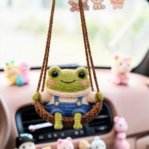 May include: A crocheted green frog wearing a sweater and blue overalls, sitting in a brown swing. The frog is hanging from a car's rearview mirror. Decorative figurines of bears and other characters are also visible.