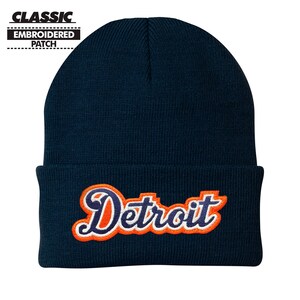 May include: A navy blue beanie with a white and orange embroidered patch that says "Detroit" in a retro script font.