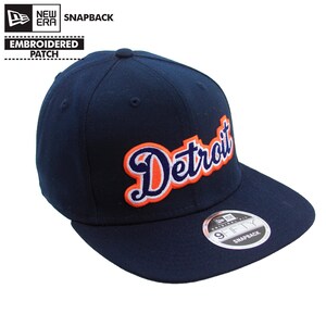 May include: A navy blue snapback baseball cap with the word "Detroit" embroidered in orange and white script. The cap features a curved bill and a "9FIFTY" sticker. The New Era logo is visible.
