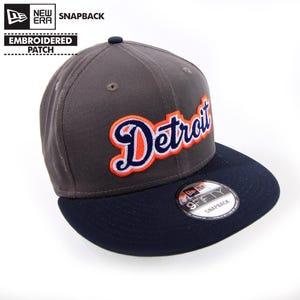 May include: A gray and navy blue New Era 9FIFTY snapback hat with a white and orange embroidered "Detroit" logo.