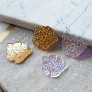 Puede incluir: Cuatro abalorios de mariposa de acrílico con acabado dorado e iridiscente. Las mariposas tienen un diseño detallado con una luna y estrellas.