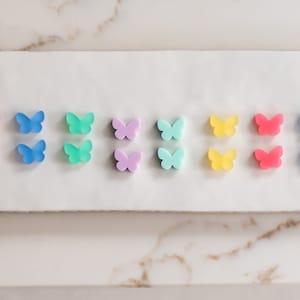 May include: A set of 10 butterfly-shaped earrings in various pastel colors, including pink, blue, green, yellow, and purple. The earrings are arranged in pairs on a white surface.