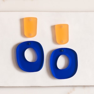 Puede incluir: Un par de pendientes de resina azul con forma ovalada redondeada y un recorte ovalado más pequeño en el centro. Los pendientes se emparejan con dos pendientes de resina naranja más pequeños.