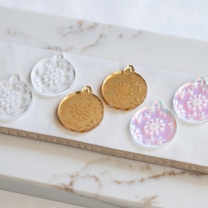May include: Five round, clear acrylic ornaments with snowflake designs. Two ornaments are gold, two are iridescent, and one is clear.