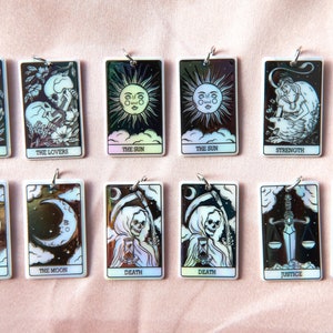 Puede incluir: Doce amuletos de cartas de tarot rectangulares blancas iridiscentes con ilustraciones en blanco y negro y anillos plateados. Las cartas incluyen Los Enamorados, El Sol, La Fuerza, La Luna, La Muerte y La Justicia.