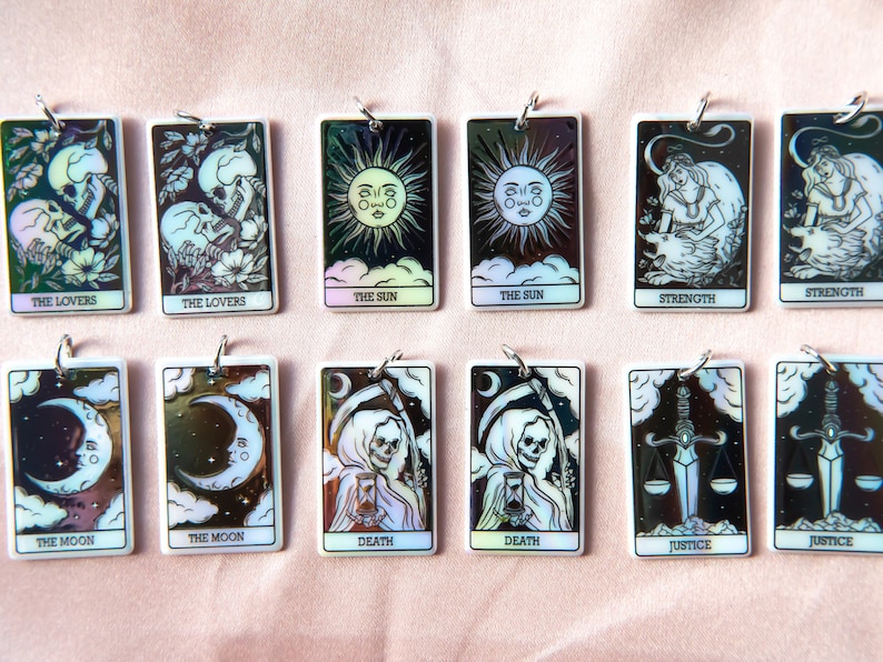 Puede incluir: Doce amuletos de cartas de tarot rectangulares blancas iridiscentes con ilustraciones en blanco y negro y anillos plateados. Las cartas incluyen Los Enamorados, El Sol, La Fuerza, La Luna, La Muerte y La Justicia.