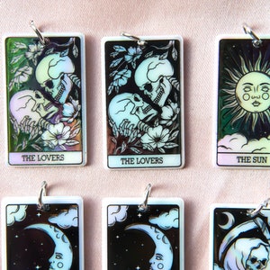 Puede incluir: Seis amuletos de cartas de tarot iridiscentes con anillos de plata. Las cartas son Los Enamorados, Los Enamorados, El Sol, La Luna, La Luna y la Muerte. Las cartas est&aacute;n sobre un fondo rosa.