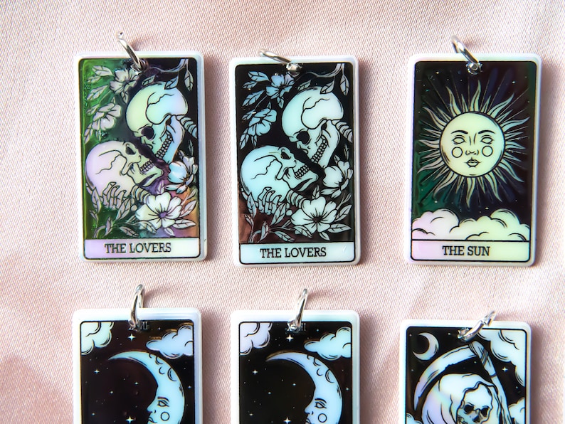 Puede incluir: Seis amuletos de cartas de tarot iridiscentes con anillos de plata. Las cartas son Los Enamorados, Los Enamorados, El Sol, La Luna, La Luna y la Muerte. Las cartas est&aacute;n sobre un fondo rosa.