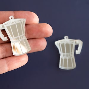 May include: Two clear, mirrored coffee pot charms. One charm is held in a hand, while the other sits on a dark blue surface. The charms are made of a clear material with a reflective, mirrored interior, and a small loop at the top.