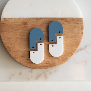 Op de afbeelding: Twee paar oorbellen, een blauwe en een witte, met een geometrische vorm. De oorbellen liggen op een houten oppervlak.
