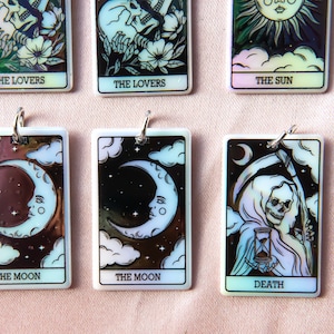 Puede incluir: Un conjunto de diez amuletos de cartas de tarot iridiscentes con im&aacute;genes de Los Enamorados, El Sol, La Luna y La Muerte. Cada amuleto es un colgante rectangular con una imagen en blanco y negro sobre un fondo blanco.