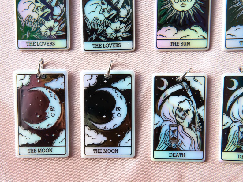 Puede incluir: Un conjunto de diez amuletos de cartas de tarot iridiscentes con im&aacute;genes de Los Enamorados, El Sol, La Luna y La Muerte. Cada amuleto es un colgante rectangular con una imagen en blanco y negro sobre un fondo blanco.