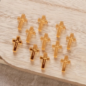 May include: A collection of twelve gold-colored cross charms, each with a raised design, arranged on a light wood surface.
