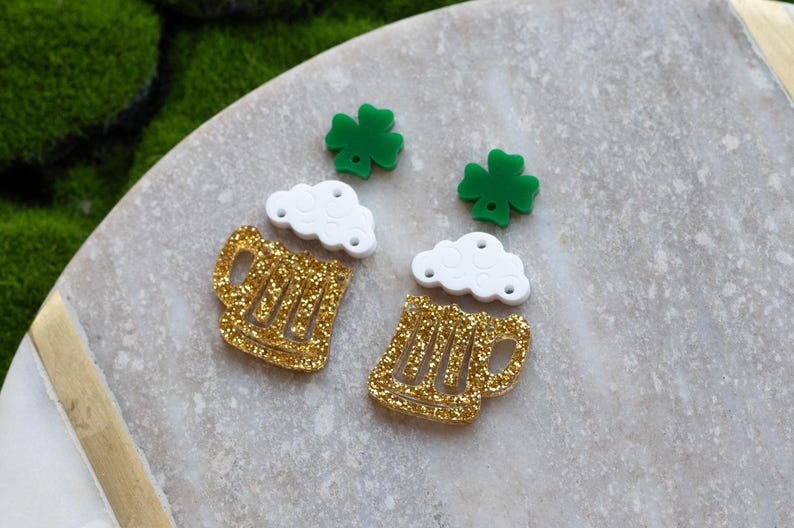 May include: A pair of gold glitter beer mug earrings with white foam and green shamrock charms.