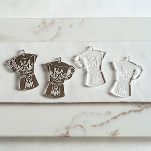 May include: Four silver-colored metal earrings shaped like coffee pots with a floral design. The earrings are on a white surface.