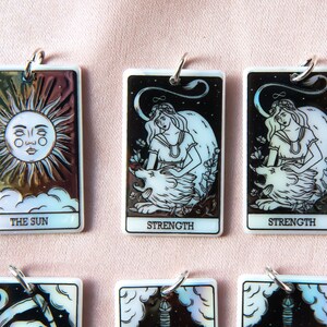 Puede incluir: Un conjunto de seis amuletos de cartas de tarot iridiscentes con las cartas Fuerza, Sol y Luna. Cada amuleto es rectangular y tiene un anillo plateado para colgar.