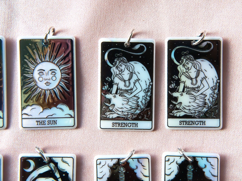 Puede incluir: Un conjunto de seis amuletos de cartas de tarot iridiscentes con las cartas Fuerza, Sol y Luna. Cada amuleto es rectangular y tiene un anillo plateado para colgar.