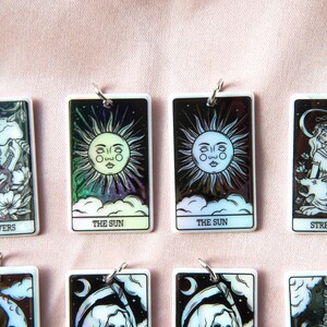 Puede incluir: Ocho cartas de tarot rectangulares blancas iridiscentes con ilustraciones en blanco y negro y anillos plateados. Las cartas representan Los Enamorados, El Sol, La Fuerza, La Luna, La Muerte y La Torre.