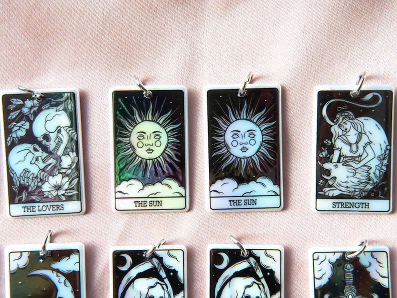 Puede incluir: Ocho cartas de tarot rectangulares blancas iridiscentes con ilustraciones en blanco y negro y anillos plateados. Las cartas representan Los Enamorados, El Sol, La Fuerza, La Luna, La Muerte y La Torre.