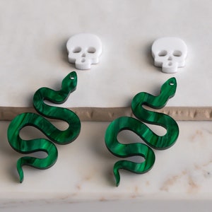 Puede incluir: Un par de pendientes de serpiente verde con un dije de calavera blanca. Los pendientes de serpiente están hechos de un material verde que se asemeja a la malaquita.