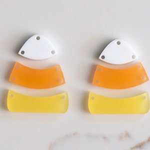May include: Two sets of candy corn-shaped acrylic charms. Each set includes a white, orange, and yellow charm. The charms have holes for attaching to jewelry or other crafts.