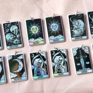 Puede incluir: Un conjunto de 12 amuletos de cartas de tarot iridiscentes con ilustraciones en blanco y negro de las cartas de los Arcanos Mayores. Los amuletos est&aacute;n hechos de resina blanca y tienen un lazo de metal plateado para colgar.