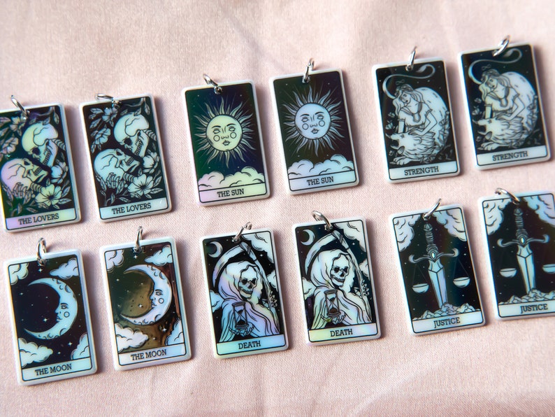 Puede incluir: Un conjunto de 12 amuletos de cartas de tarot iridiscentes con ilustraciones en blanco y negro de las cartas de los Arcanos Mayores. Los amuletos est&aacute;n hechos de resina blanca y tienen un lazo de metal plateado para colgar.