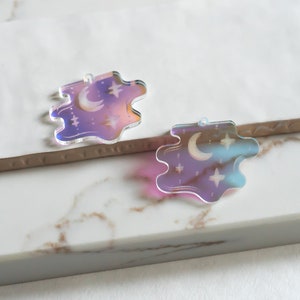 May include: Two iridescent acrylic charms shaped like a cloud with a crescent moon and stars. The charms are a light purple and blue color.
