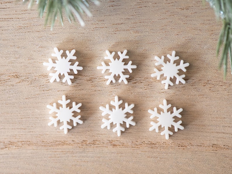 Pu&ograve; includere: Sei ornamenti bianchi a forma di fiocco di neve su una superficie di legno marrone chiaro.