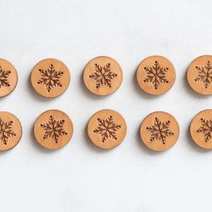May include: Ten wooden round ornaments with a snowflake design. The ornaments are light brown and have a natural wood finish.