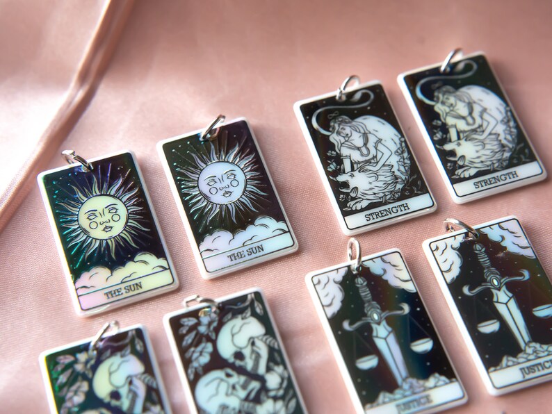 Puede incluir: Un conjunto de ocho amuletos de cartas del tarot iridiscentes que presentan varias im&aacute;genes de cartas del tarot, incluyendo El Sol, la Fuerza y ​​la Justicia. Los amuletos est&aacute;n hechos de acr&iacute;lico blanco y tienen un acabado hologr&aacute;fico.