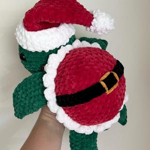 May include: A handmade green and red plush turtle wearing a Santa hat and belt. The turtle's shell is red with a black belt and gold buckle. The hat has a white trim and pom-pom. A festive holiday decoration.