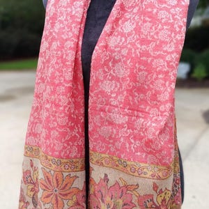 May include: A pink and beige patterned scarf with a floral design. The scarf has a border with a floral pattern in shades of pink, orange, and green.