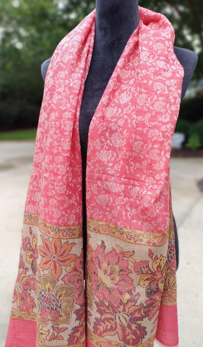 May include: A pink and beige patterned scarf with a floral design. The scarf has a border with a floral pattern in shades of pink, orange, and green.