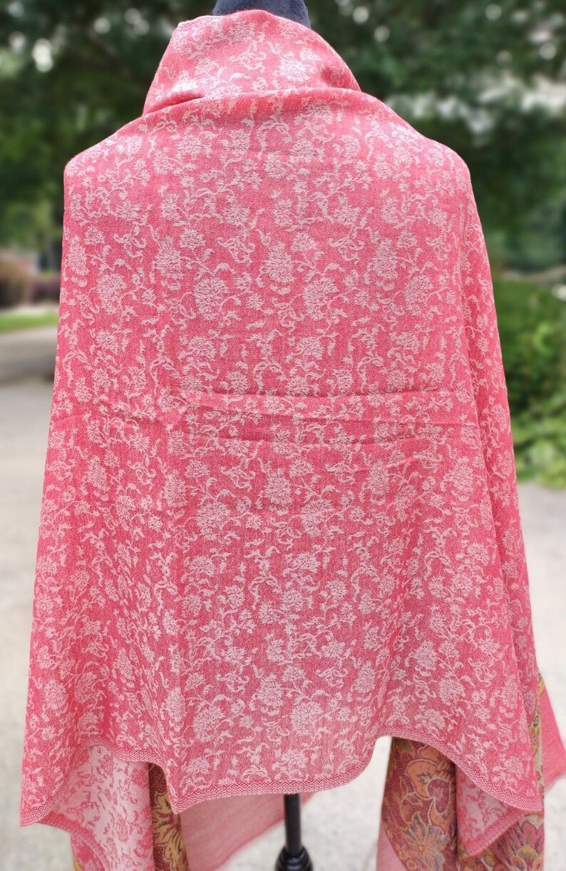 May include: A pink shawl with a white floral pattern. The shawl is draped over a mannequin and has a fringed edge.