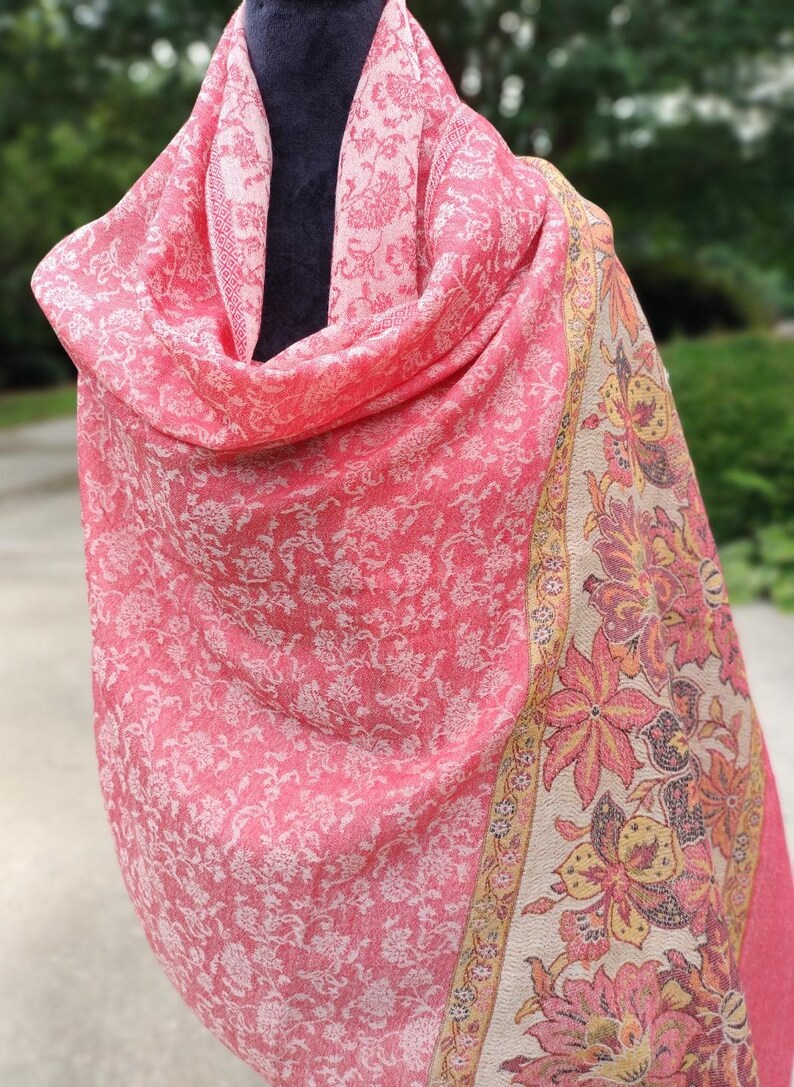 May include: A pink shawl with a white floral pattern and a beige border with a floral design. The shawl is draped over a mannequin.